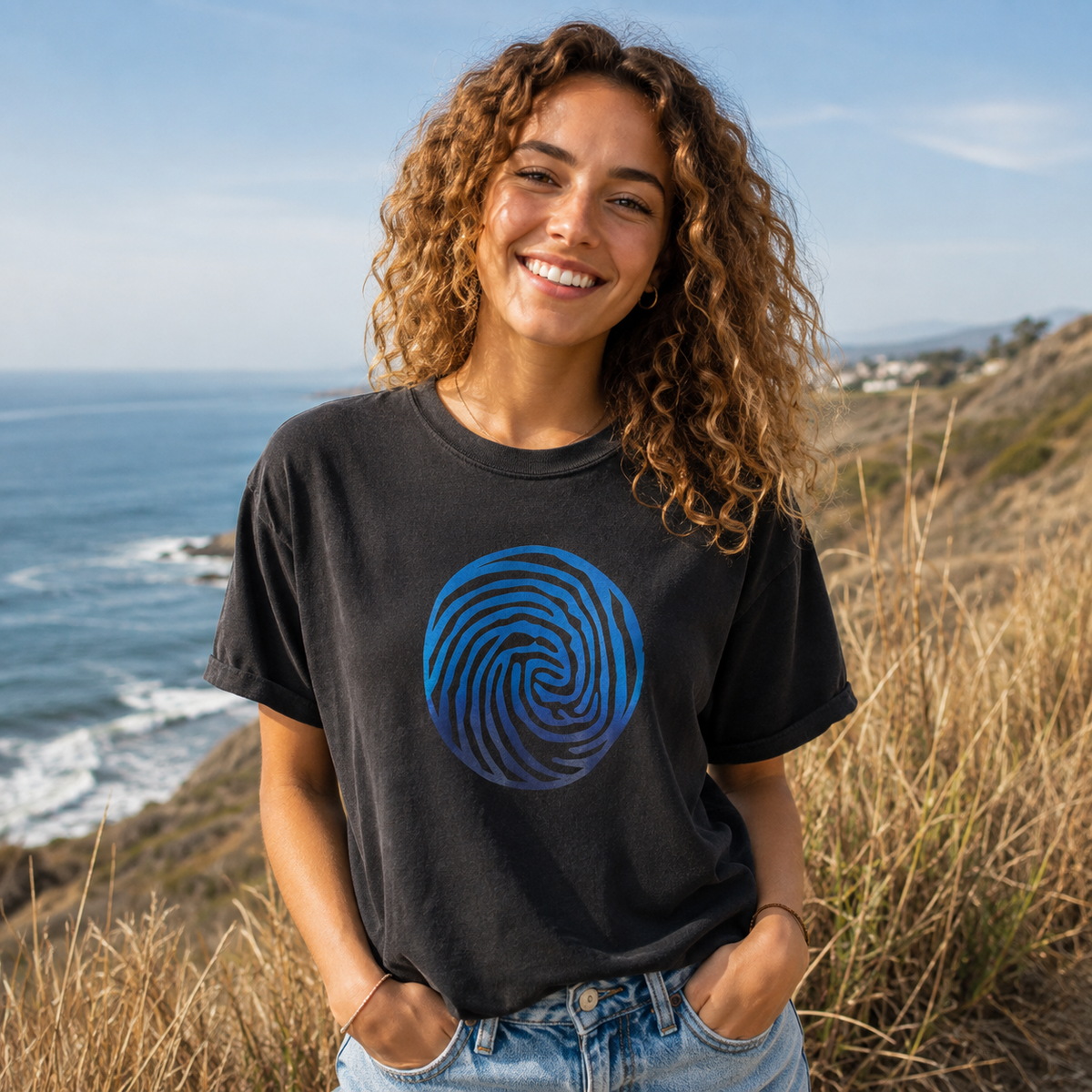 A smiling woman with curly hair stands outdoors on a grassy cliffside overlooking the ocean, wearing a faded black graphic T-shirt with a blue spiral design on the front and light-wash jeans.
