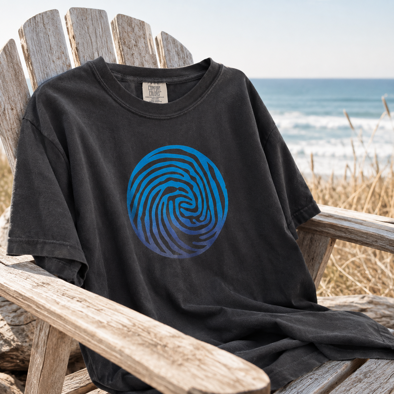 A black short-sleeve t-shirt with a blue spiral fingerprint graphic on the front, displayed on a weathered wooden beach chair with the ocean in the background.