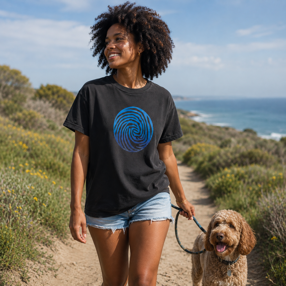 A woman walking a brown dog on a dirt path outdoors, wearing a faded black t-shirt with a blue spiral graphic on the front. The background shows a coastal landscape with the ocean, sky, and green shrubs.
