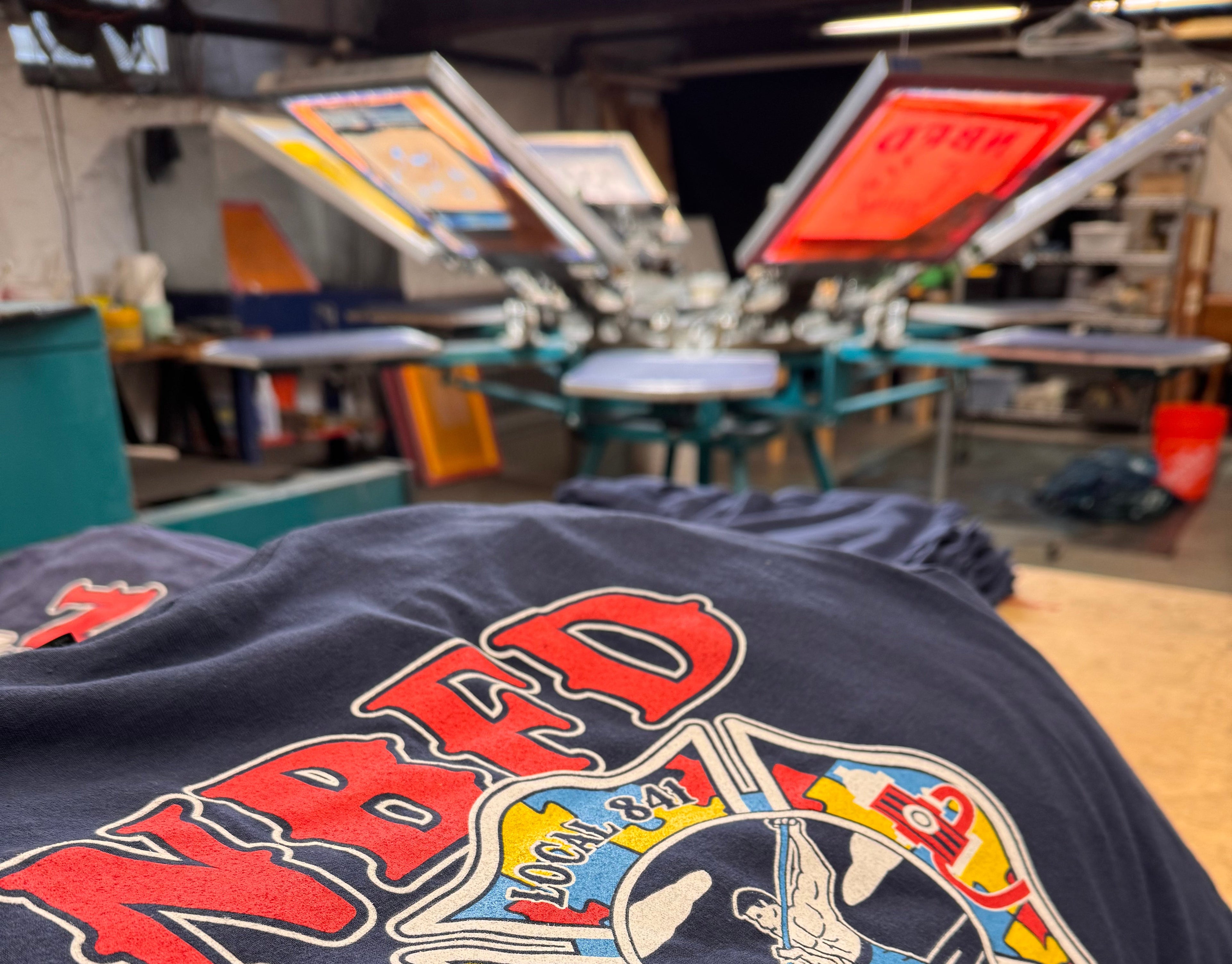 Close-up of freshly screen printed navy shirts on a print shop table with a multi-station screen printing press in the background at Crazy Fingers Design.
