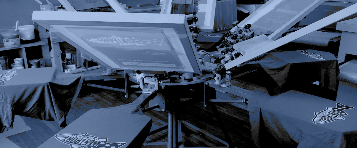 A large industrial screen printing press with a large frame holding a printed sheet, surrounded by tables covered in matching printed cloths at what appears to be a trade show or workshop.