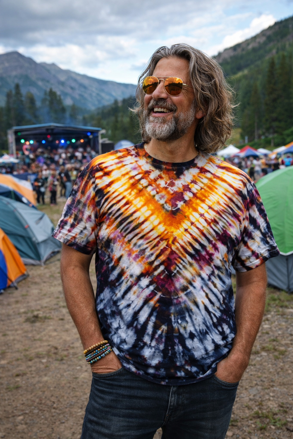 Man wearing a tie-dye shirt at a music festival with tents and mountains in the background
